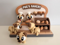A miniature toy bakery display featuring cartoon pug dogs dressed as bakers and several small breads on shelves, with a sign reading 'PUG'S BAKERY' at the top.