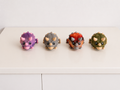 Four small dinosaur figurines lined up on a white surface against a white wall. From left to right, they are purple with pink horns, gray with tan horns, orange with red horns, and olive green with tan horns.